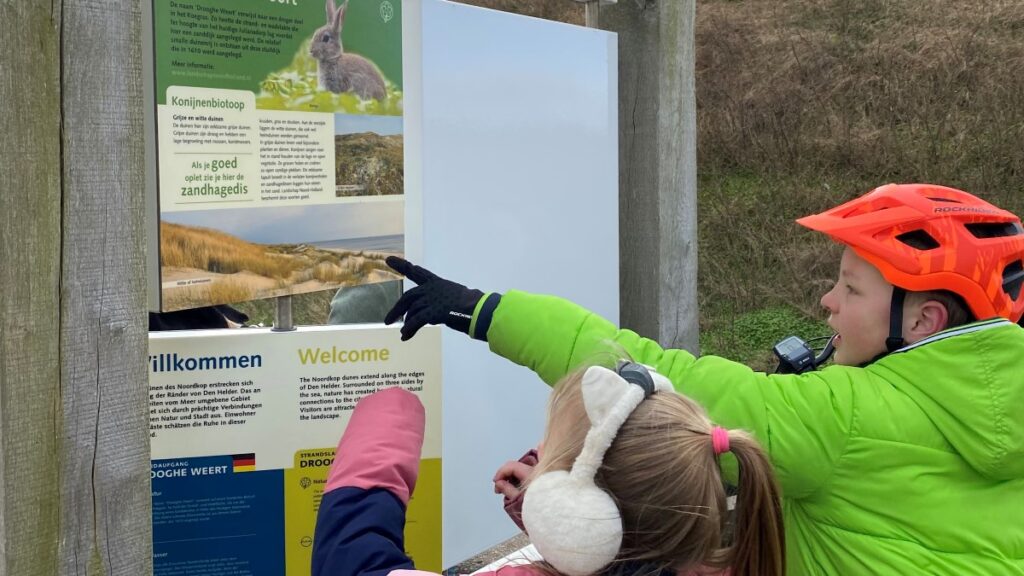 De kinderen kijken op het bord bij de ingang van de duinen om een aanwijzing te ontdekken van Puzzelen op de Pedalen.